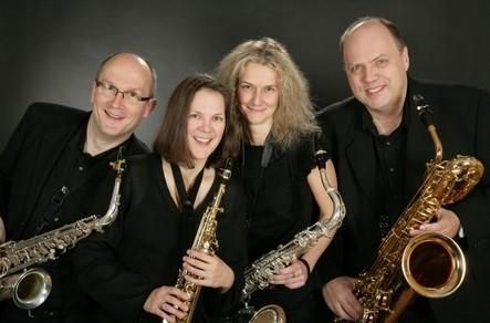 Eine Jazzband mit vier Musikern steht gemeinsam in Schwarz gekleidet da. Jeder hält ein Saxophon und lächelt in die Kamera.