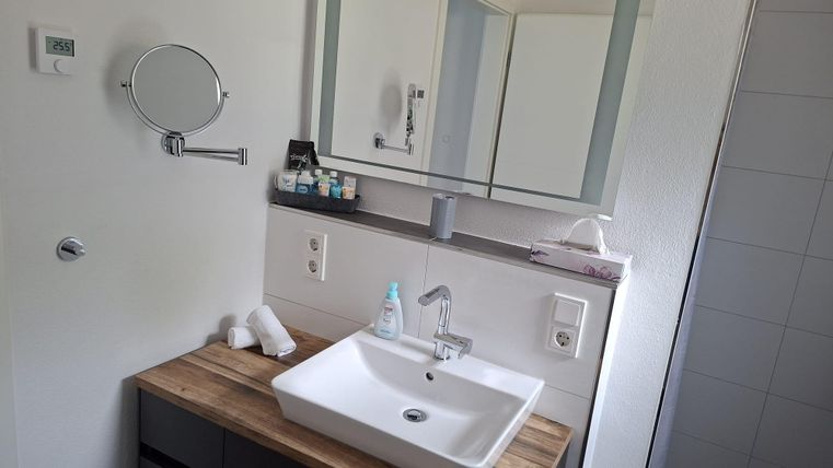Ein modernes Badezimmer mit einem Waschbecken auf einer Holzablage. Im Hintergrund sind Handtücher und verschiedene Pflegeprodukte zu sehen.