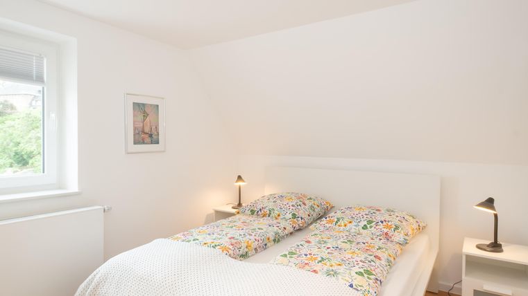 Ein helles Schlafzimmer mit einem Doppelbett und bunten Bettdecken. An der Wand hängt ein Bild, und es gibt zwei Nachttischlampen.