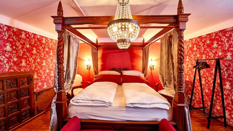 Ein elegantes Schlafzimmer mit einem Himmelbett und einer großen Deckenleuchte. Die Wände sind mit auffälligem rotem Blumentapete dekoriert.