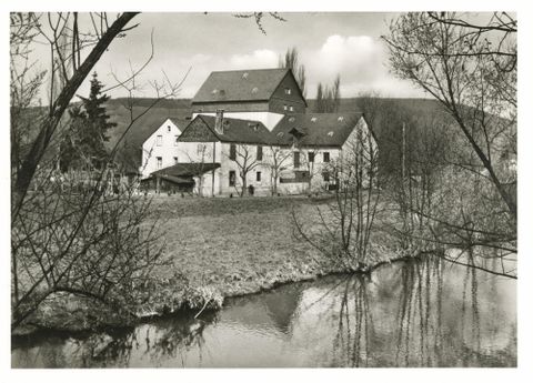 Een historisch gebouw aan een rustig water, omgeven door bomen. De scène is in zwart-wit en roept een nostalgische sfeer op.