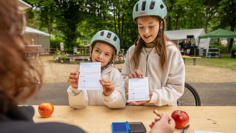 Twee meisjes met fietshelmen houden kaarten in de camera. Op een tafel liggen appels en een klein doosje.