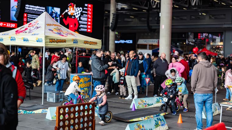 Een levendige gebeurtenis met veel mensen en kinderen die op kleine fietsen rijden. Op de achtergrond zijn tenten en een hoop activiteiten te zien.