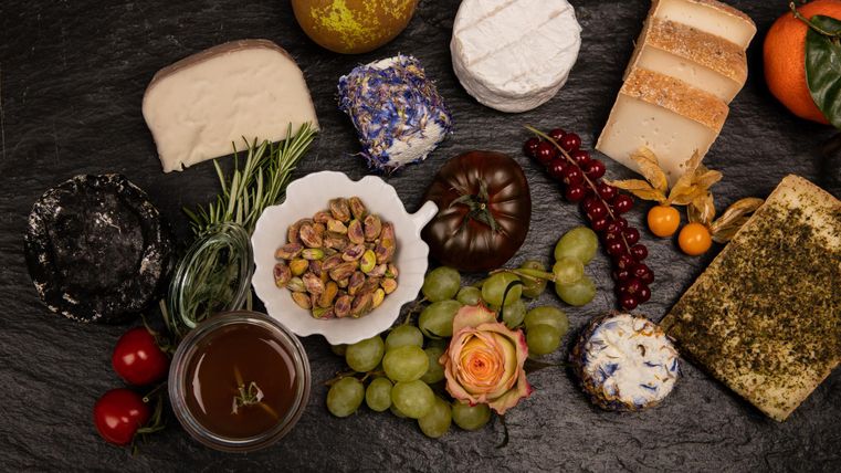 A selection of various cheeses, fresh fruits, and nuts on a black background. The arrangement also includes bread and edible flowers.