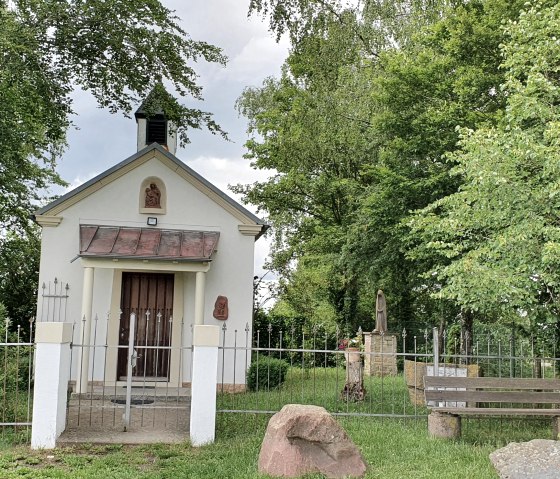 Een kleine witte kapel met een hek omringd door bomen. Een bank en stenen staan op de voorgrond., © TI Bitburger Land