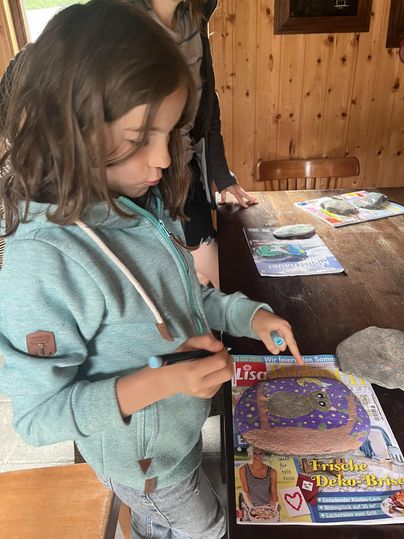 Un enfant dessine sur un magazine avec un stylo. En arrière-plan, on peut voir d'autres magazines et une table en bois.