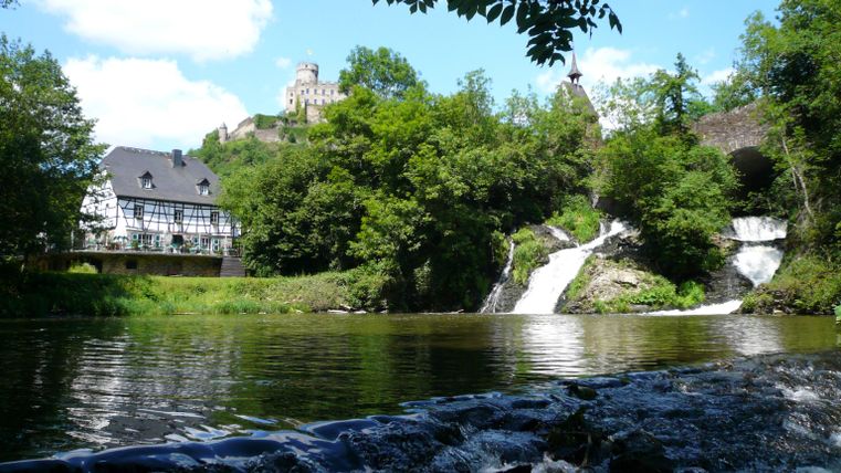A picturesque waterfall flows into a tranquil river. In the background, trees and a castle can be seen under a clear blue sky.