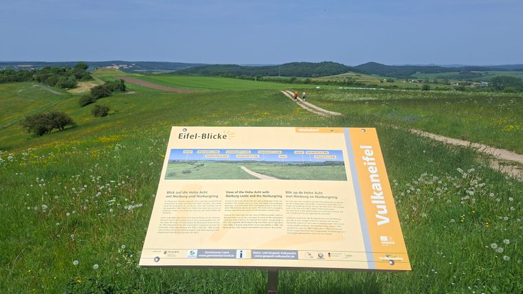 Un point de vue avec un panneau d'information sur l'Eifel. Le paysage est vert et vallonné sous un ciel bleu clair.