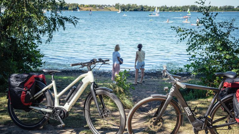 Two bicycles stand on the shore of the Zülpich water sports lake, sailing boats can be seen on the water in the background.
