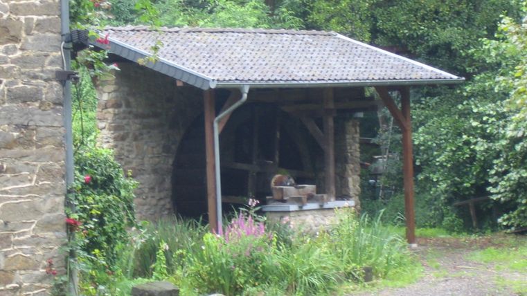 An old mill house with a water wheel and green undergrowth. The surroundings are quiet and surrounded by trees.