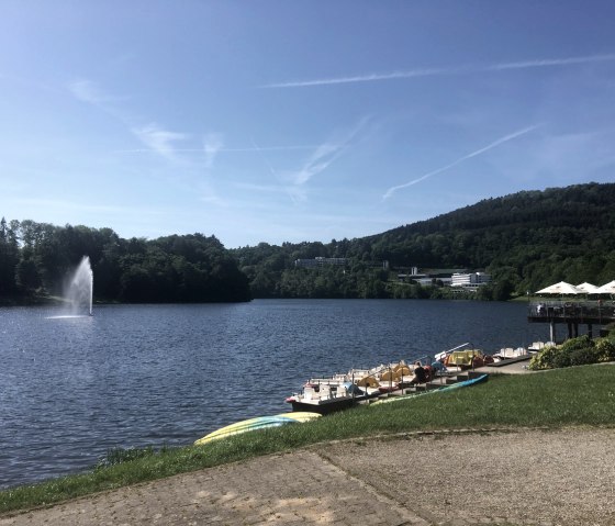 The Bitburg reservoir in Biersdorf am See with pedal boats, a fountain and surrounding hills under a blue sky., © TI Bitburger Land