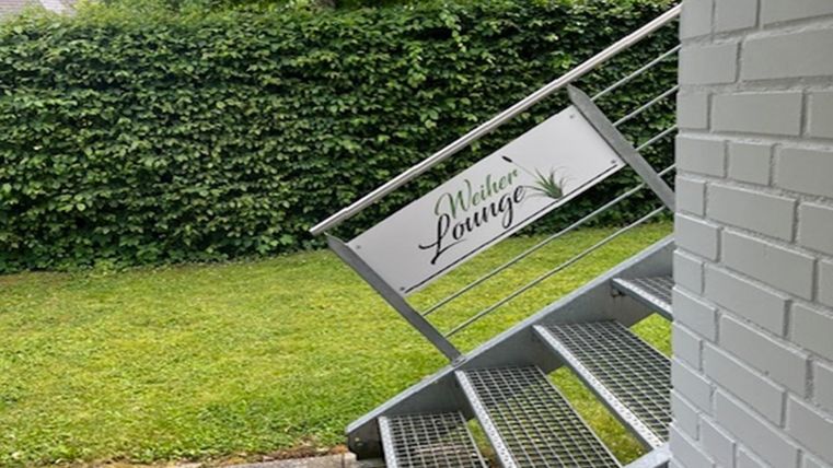 Une vue extérieure avec un escalier en métal et un panneau indiquant la "Welker Lounge". En arrière-plan, on peut voir des buissons verts et une pelouse bien entretenue.