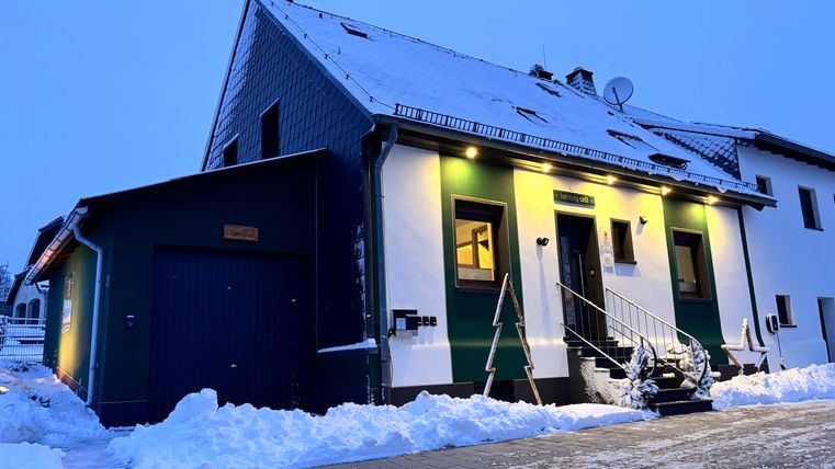 A pretty house with a white and green façade, surrounded by snow. It is evening and the windows are brightly lit.