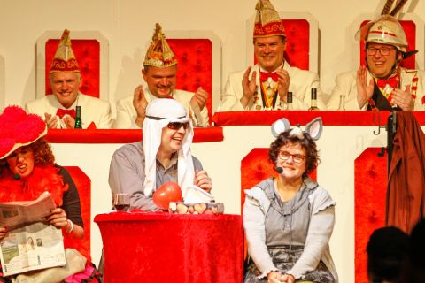 A group of people in colorful costumes sits on a stage. Some are applauding, while others are seated at a table with fruit and accessories.