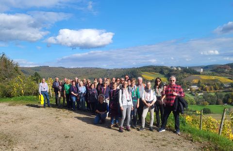 Eine Gruppe von Menschen steht auf einem Weg mit Blick auf eine malerische Landschaft. Der Himmel ist blau mit wenigen Wolken und die Umgebung ist grün und hügelig.