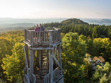 Zwei Personen stehen auf dem hölzernen Aussichtsturm Dietzenley am Keltenpfad und genießen den weiten Blick über die bewaldete Eifellandschaft bei klarer Sicht und sanftem Licht.