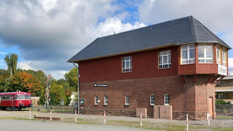 A historic building with red brickwork and a gray roof. In the foreground, there is an old bus and the sky is cloudy.