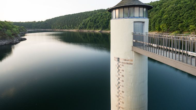 Blick auf den Urftsee mit einem Turm und Brücke im Vordergrund, umgeben von bewaldeten Hügeln.