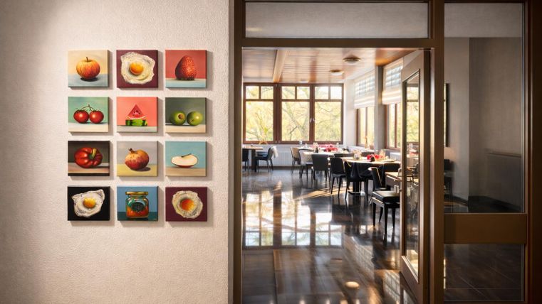 Eine moderne Restaurantatmosphäre mit gemalten Obstmotiven an der Wand. Durch die große Glasfront ist der Essbereich mit Tischen und Stühlen gut sichtbar.