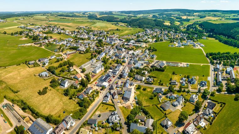 Ein weitläufiges Landschaftsbild mit einem kleinen Dorf, umgeben von grünen Feldern und Hügeln. Die Sonne scheint und der Himmel ist klar.