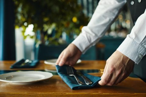 Ein Kellner deckt einen Tisch mit Besteck und einer Serviette. Der Tisch ist elegant und gut vorbereitet für Gäste.