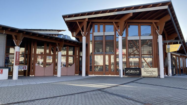 Ein modernes Gebäude mit großen Fenstern und einem modernen Holzdesign. Der Platz vor dem Gebäude ist mit Pflastersteinen ausgelegt.