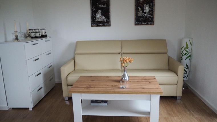 Ein gemütliches Wohnzimmer mit einem beigen Sofa und einem Holztisch. An der Wand hängen zwei Bilder, und neben dem Tisch steht eine Vase mit Blumen.