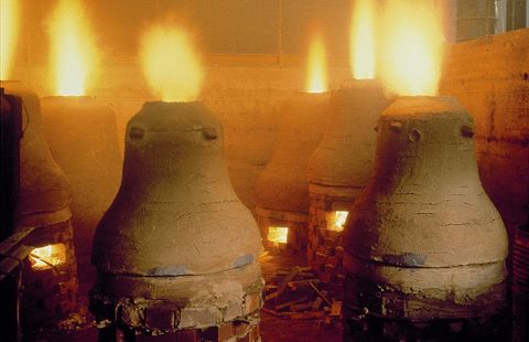 Dans une ancienne salle de four, plusieurs grands fours se dressent, d'où s'élèvent des flammes. Les murs sont en pierre et l'atmosphère est chaude et fumée.