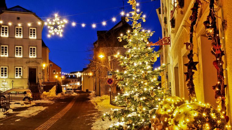 Une rue décorée de manière festive avec un arbre de Noël illuminé et des lumières. Le ciel nocturne est clair et la neige recouvre le sol.