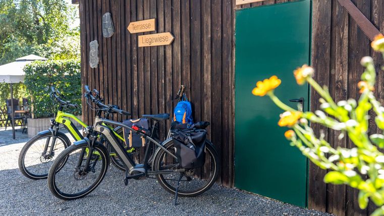A wooden bicycle garage with two bicycles and a blue backpack. In the background, plants and an outdoor table can be seen.