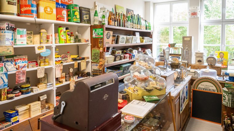 Un magasin confortable et nostalgique avec des étagères remplies d'emballages et de produits d'autres temps. Au premier plan se trouve une vieille caisse et il y a de nombreux articles différents à découvrir.