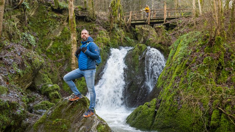 Randonneur avec sac à dos sur un rocher devant la cascade "Die Rausch" dans la vallée de la Wilden Endert, avec en arrière-plan un pont en bois au-dessus des gorges couvertes de mousse.