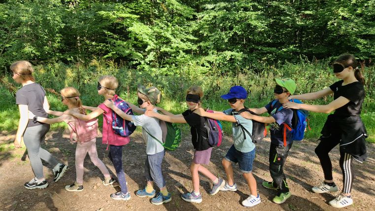A group of children is walking in the forest while wearing blindfolds. They are equipped with backpacks and are holding on to each other's shoulders.