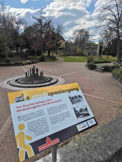 Een park met een fontein en een informatiebord over een stadswandeling.