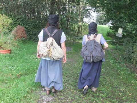 Two women with backpacks are walking along a narrow path. Surrounded by trees and green grass, the scene appears calm and connected to nature.