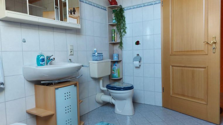 Een heldere badkamer met witte tegels en een houten kast. Het heeft een wastafel, een toilet en een blauwe tapijt.