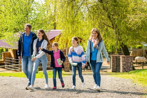 Eine Familie mit zwei Erwachsenen und drei Kindern läuft fröhlich durch einen Wildpark. Im Hintergrund stehen Bäume, Holzhütten und Gehege. Die Sonne scheint und alle tragen bunte Kleidung.