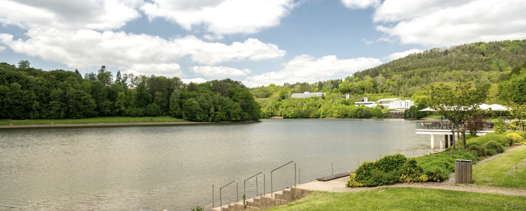 Stausee Bitburg mit grüner Uferlandschaft, Bäumen und Gebäuden im Hintergrund unter einem bewölkten Himmel., © Eifel Tourismus GmbH, Dominik Ketz