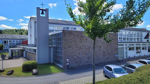 A modern building with a church tower bell and large windows. In the foreground, we see some cars and trees.