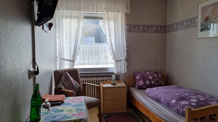 Ein einfaches Schlafzimmer mit einem Einzelbett, einem Nachttisch und einem Fernseher. Das Fenster ist mit Vorhängen dekoriert und es gibt einen Tisch mit einer Flasche Wasser.