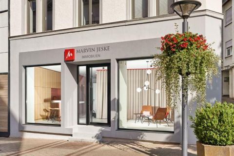 Ein modernes Immobilienbüro mit großen Fenstern und klaren Linien. Die Fassade ist hell und einladend, umgeben von grünen Pflanzen.