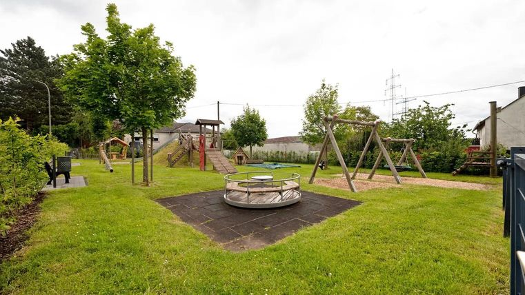 Ein Spielplatz mit verschiedenen Spielgeräten und viel grünem Gras. Umgeben von Bäumen und einem ruhigen, grauen Himmel.