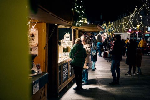 Een drukke kerstmarkt 's nachts met feestelijke verlichting. Mensen snuffelen aan houten kraampjes met waren.