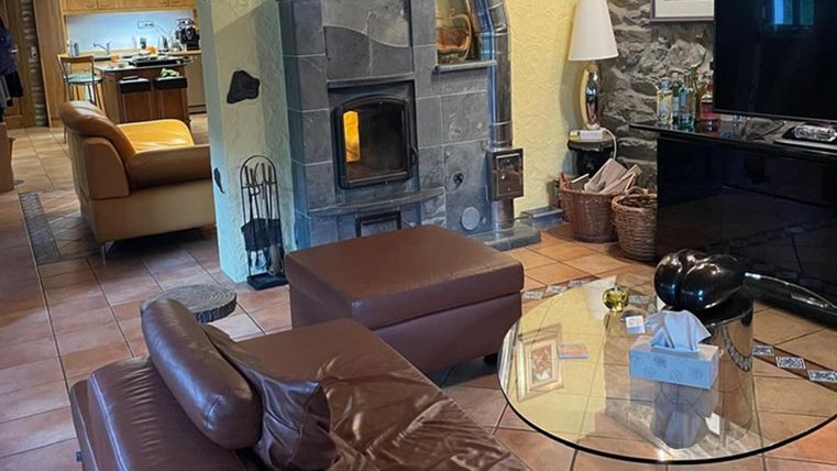 A cozy living room with a dark brown sofa and a round glass table. In the background, there is an open space with a modern kitchen and a wood stove.