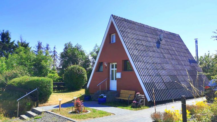 Ein gemütliches, rot getünchtes Ferienhaus mit einem Satteldach. Umgeben von grünen Büschen und einem klaren blauen Himmel.