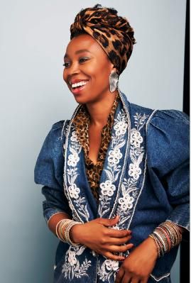 A cheerful woman with a striking headscarf and a beautiful top. She is wearing many colorful bracelets and smiling warmly at the camera.