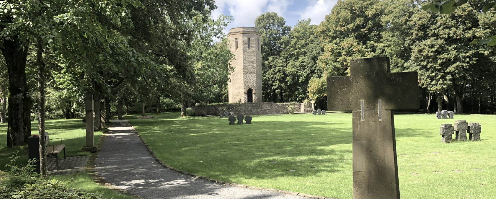 Ein Friedhof mit steinernen Kreuzen, einem Turm im Hintergrund und einem Weg, der von Bäumen gesäumt ist. Der Himmel ist teils bewölkt.