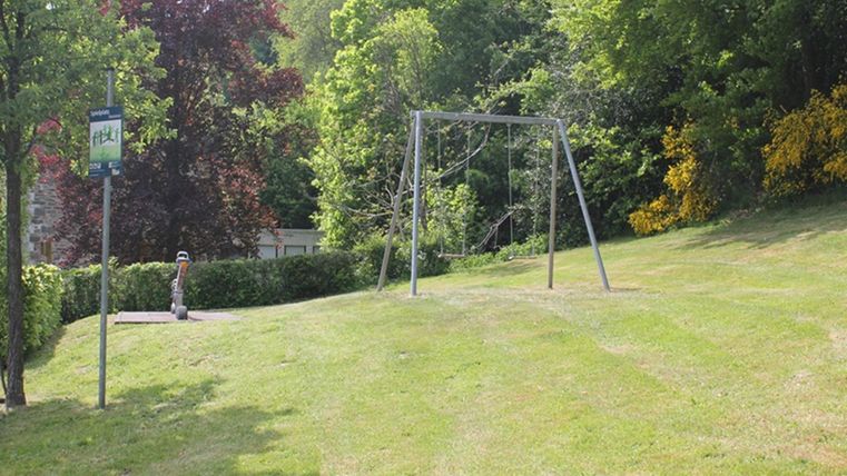 Een rustige park met een schommel en weelderig, groen gras. Op de achtergrond zijn bomen en een bushalte te zien.