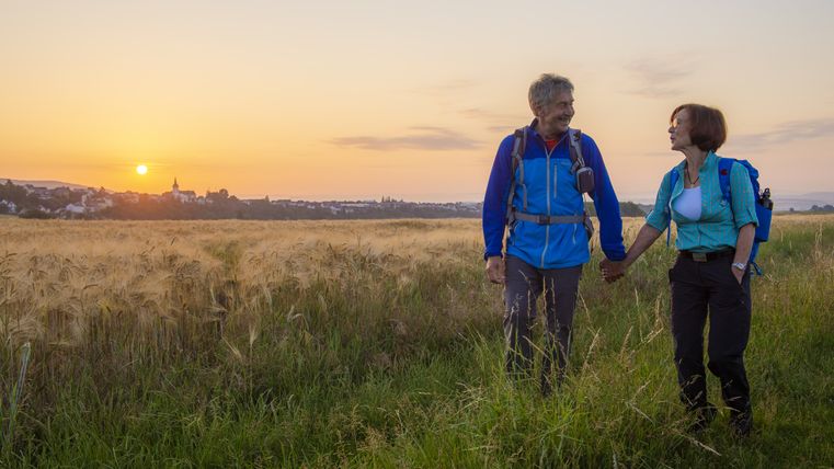 Een koppel wandelt door een veld bij zonsondergang, met een stad op de achtergrond.