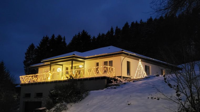 Een gezellig huis met feestelijke verlichting in een besneeuwd landschap. Op de voorgrond staat een versierde kerstboom.